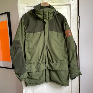 Könnte beinhalten: Eine grüne und olivfarbene Kapuzenjacke mit mehreren Taschen und einem Aufnäher am oberen rechten Ärmel. Die Jacke hat einen Reißverschluss vorne und einen hohen Kragen. Die Jacke hängt an einem Holzbügel.