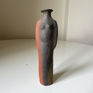 Op de afbeelding: Een handgemaakte keramische vaas met een unieke, mensachtige vorm. De vaas heeft een donkerbruine en rood-oranje kleurovergang, met een getextureerd oppervlak. Het ontwerp omvat een uitlopende hals en subtiele details die een torso suggereren.