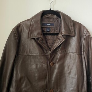 May include: A brown leather jacket with a button-up front and a collared neckline. The jacket has a quilted lining and the brand name "GAP" is visible on a label. The jacket is hanging on a black hanger.