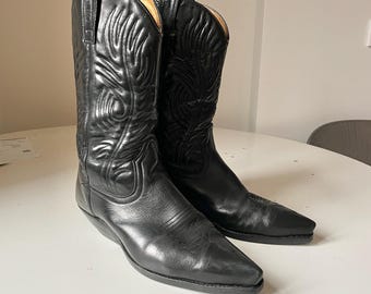 May include: A pair of black leather cowboy boots featuring a pointed toe, a stacked heel, and intricate decorative stitching patterns on the tall shaft. The boots have pull tabs at the top and a smooth, polished finish throughout the exterior.