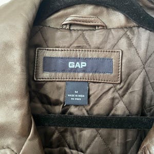 May include: Close-up of a brown leather jacket with a quilted lining. The jacket has a collar and a label that reads "GAP". The label also indicates the size is medium and "Made in India".