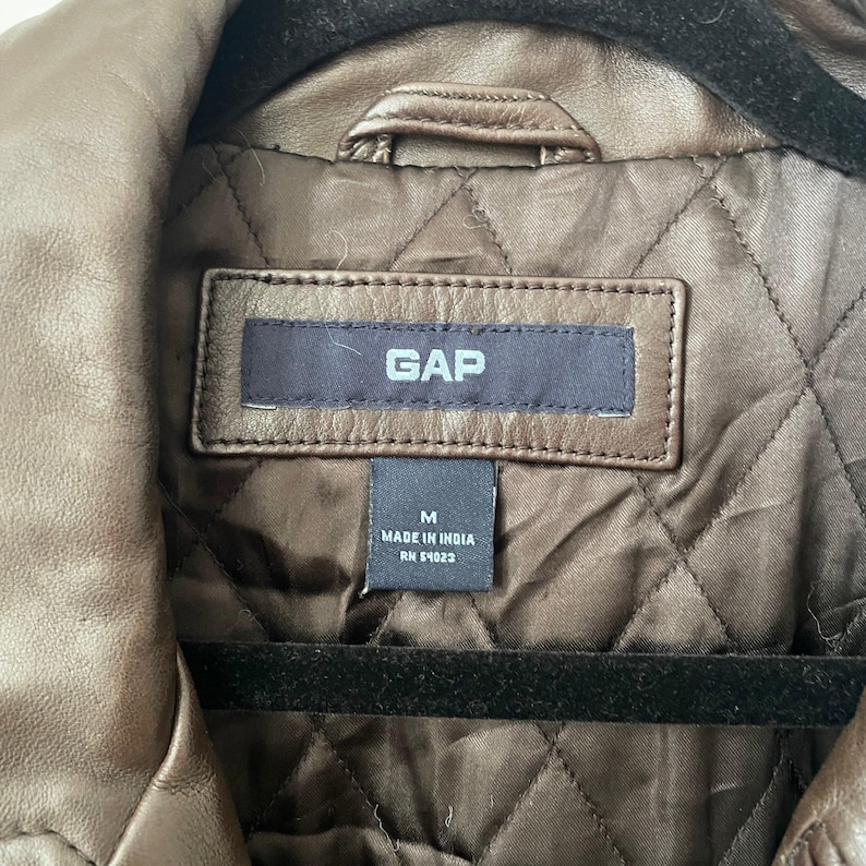 May include: Close-up of a brown leather jacket with a quilted lining. The jacket has a collar and a label that reads "GAP". The label also indicates the size is medium and "Made in India".