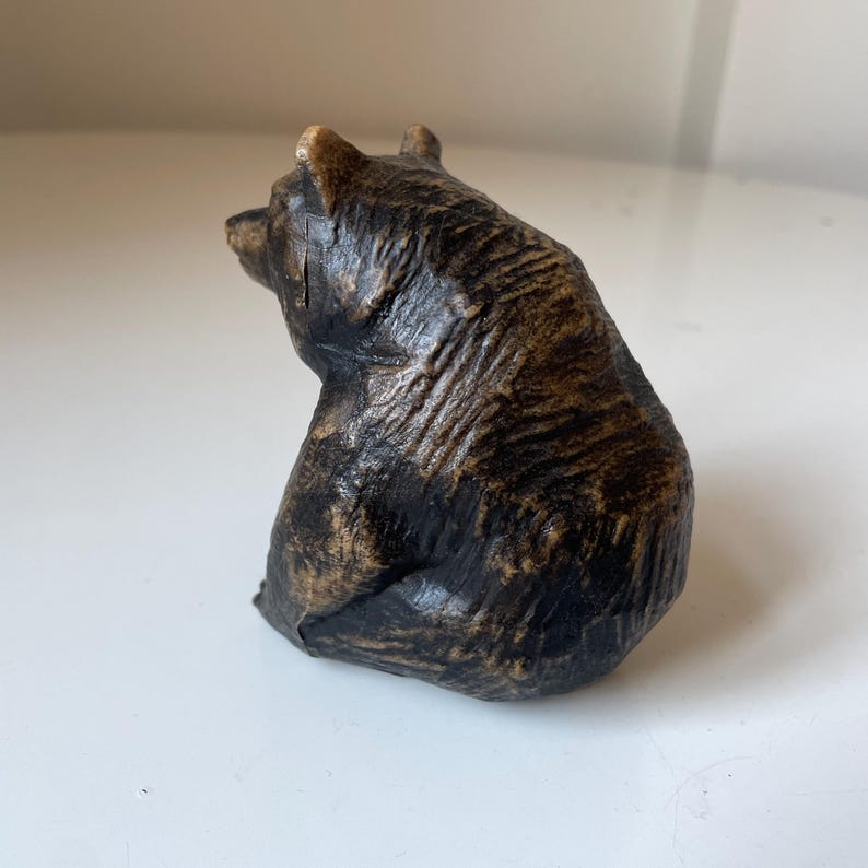 May include: A small, detailed bear figurine in shades of brown and black. The bear is sitting, with its back to the viewer, showcasing textured fur and a rustic appearance. This decorative item could be a collectible or a unique home decor piece.