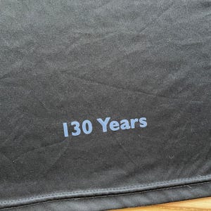 May include: Close-up of a black fabric with the text "130 Years" printed in light blue. The fabric has a slightly textured appearance and a stitched hem at the bottom. The text is centered and clearly visible.