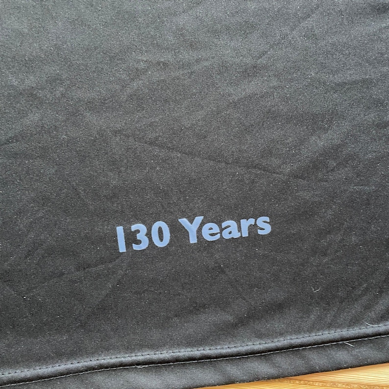 May include: Close-up of a black fabric with the text "130 Years" printed in light blue. The fabric has a slightly textured appearance and a stitched hem at the bottom. The text is centered and clearly visible.