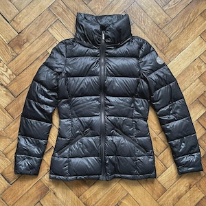 Könnte beinhalten: Schwarze Steppjacke mit hohem Kragen und durchgehendem Reißverschluss. Die Jacke hat ein gestepptes Design und zwei Vordertaschen. Ein kleines Logo ist auf dem linken oberen Ärmel sichtbar. Für kaltes Wetter.