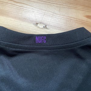 Puede incluir: Primer plano del cuello de una camiseta deportiva negra con las letras "NUFC" bordadas en morado. La camiseta est&aacute; hecha de una tela oscura y lisa.