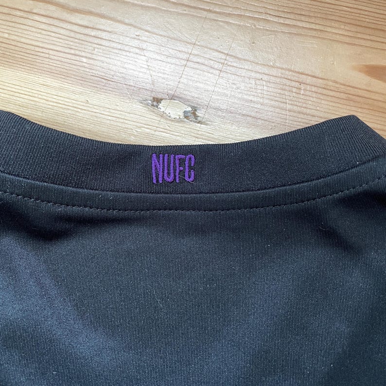 Puede incluir: Primer plano del cuello de una camiseta deportiva negra con las letras "NUFC" bordadas en morado. La camiseta est&aacute; hecha de una tela oscura y lisa.