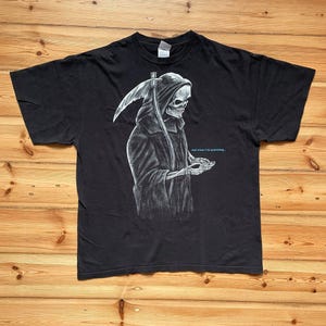 Puede incluir: Una camiseta negra de manga corta con una ilustración gráfica blanca de la muerte sosteniendo una guadaña y mirando un dispositivo portátil. El texto "not now I'm working..." está impreso en letras azules pequeñas junto a la figura.