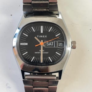 Könnte beinhalten: Eine Vintage-Timex-Uhr mit schwarzem Zifferblatt, silbernem Gehäuse und Metallarmband. Das Zifferblatt zeigt den Tag, das Datum und das Wort "Timex". Die Zeiger sind orange und die Uhr ist wasserdicht.
