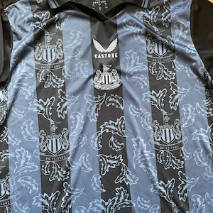 May include: A Newcastle United soccer jersey with a black and light blue striped design. The jersey features the Castore logo, the Newcastle United crest, and the date 09/12/1892. The jersey has a V-neck collar and a leaf pattern.