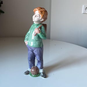 Puede incluir: Una figura de cerámica de un niño con cabello rojo, vistiendo una camisa verde de manga larga y pantalones morados. Sostiene un objeto marrón sobre su hombro y mira hacia arriba. Una pelota marrón descansa a sus pies.