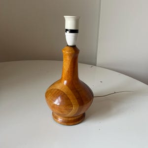 Puede incluir: Una lámpara de mesa de madera vintage con un portalámparas. La lámpara tiene una base redondeada y un cuello delgado, con un patrón de vetas de madera marrón claro y marrón oscuro. La lámpara está sobre una superficie blanca.