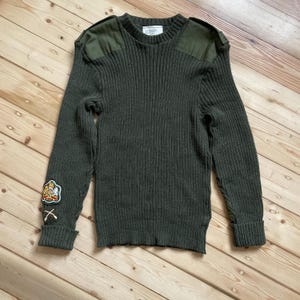 Puede incluir: Suéter de punto acanalado verde oliva con parches reforzados en los hombros y un parche en la manga izquierda. El suéter tiene cuello redondo y mangas largas. El parche presenta un diseño de escudo con herramientas cruzadas.