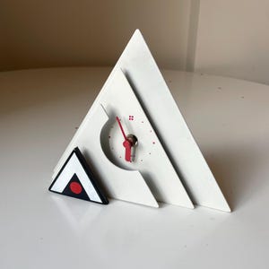 Puede incluir: Un reloj de escritorio geométrico blanco con un diseño triangular. El reloj presenta una manecilla roja y marcadores de hora de puntos rojos. Un pequeño triángulo blanco y negro con un punto rojo está al lado del reloj.