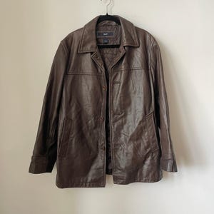 May include: A brown leather jacket with a collared neckline and button-up closure. The jacket is hanging on a black hanger. The GAP logo is visible on the label inside the jacket.