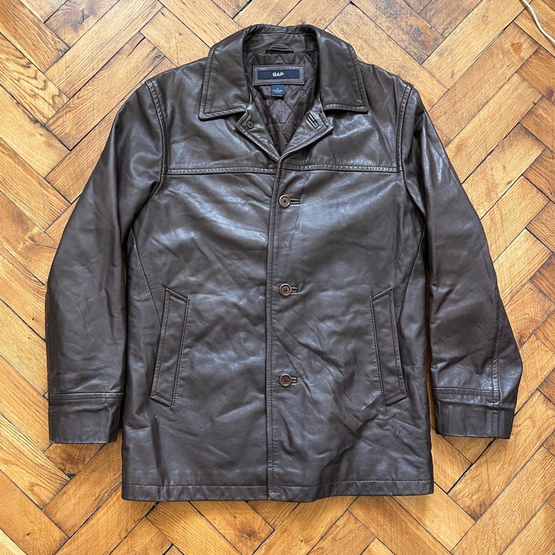 May include: A brown leather jacket with a collared neckline and button-up closure. The jacket features two front pockets and the brand name "GAP" on the label. The jacket is displayed on a wooden floor.
