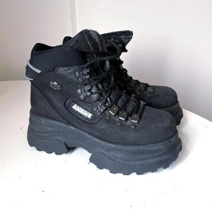 Puede incluir: Botas negras de plataforma con suela gruesa. Las botas tienen un diseño con cordones y ojales metálicos, y un cuello acolchado. La marca "Rooding's" es visible. Estas botas tienen una estética grunge o alternativa.