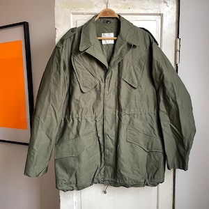 Puede incluir: Chaqueta de estilo militar verde oliva con cuello, múltiples bolsillos y cintura con cordón. La chaqueta está colgada de una percha de madera. La etiqueta de la chaqueta dice "XL".
