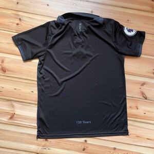 May include: Black polo shirt with a collar and short sleeves. The shirt has a logo on the sleeve and the text "NZC" on the back. The text "130 Years" is printed at the bottom. The shirt is lying on a wooden surface.