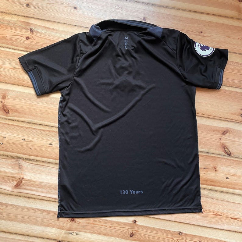 May include: Black polo shirt with a collar and short sleeves. The shirt has a logo on the sleeve and the text "NZC" on the back. The text "130 Years" is printed at the bottom. The shirt is lying on a wooden surface.