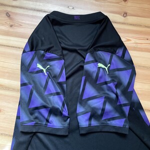 Puede incluir: Camiseta deportiva negra y morada de manga larga. Las mangas presentan un patr&oacute;n geom&eacute;trico en morado y negro, con un logotipo de Puma verde ne&oacute;n. La camiseta tiene las letras "MUFC" en la parte trasera del cuello.
