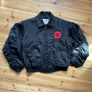 Puede incluir: Una chaqueta tipo bomber negra con cierre de cremallera frontal, cuello estándar y puños y dobladillo acanalados. La chaqueta presenta dos bolsillos delanteros con solapa y un parche circular rojo y negro en el pecho izquierdo con el texto Rammstein Lichtspielhaus.