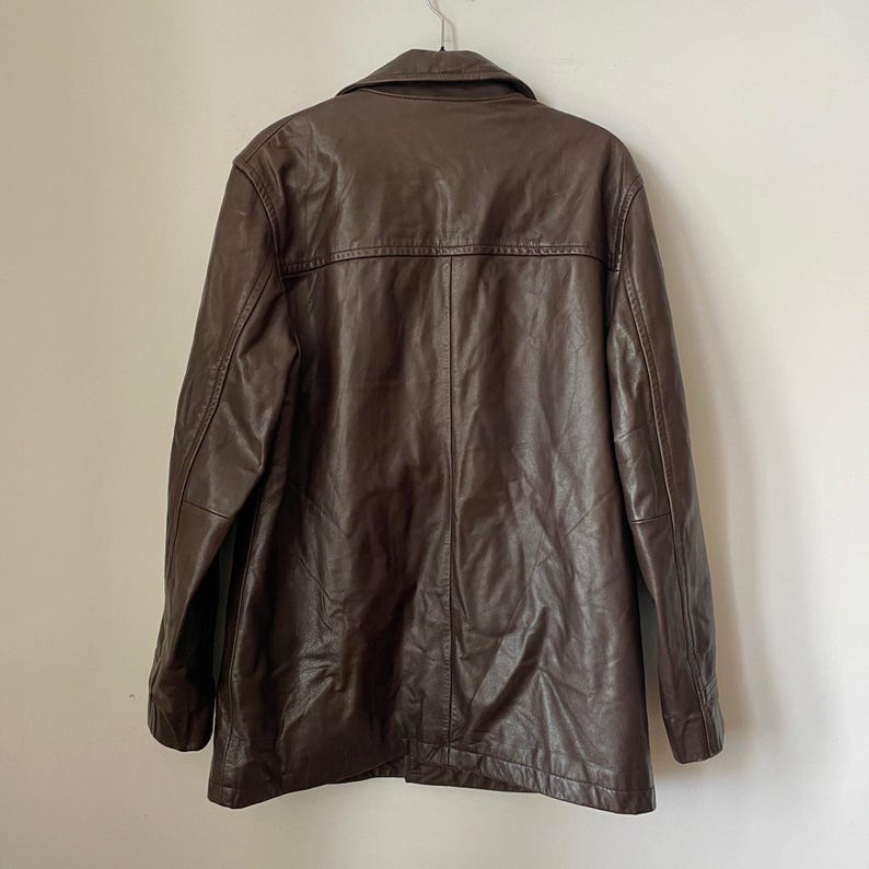 May include: A brown leather jacket hanging on a metal hanger. The jacket has a collared neckline, long sleeves, and a button-down front. The leather appears to be worn and has a slightly distressed look.