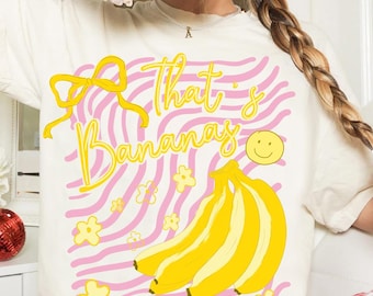 May include: Cream-colored t-shirt with a playful design. The shirt features the phrase "That's Bananas" in yellow script, a yellow bow, a bunch of bananas, a smiley face, and yellow flowers against a pink and white striped background.