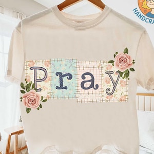 May include: Cream-colored t-shirt with the word "Pray" in a patchwork design. The letters are in a mix of floral and patterned fabrics, with pink roses and green leaves. The shirt is hanging on a wooden hanger.