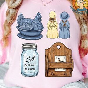 May include: A light pink sweatshirt featuring several illustrations. These include a blue hen in a bowl, two figures in dresses and bonnets, a Ball Perfect Mason jar, and a wooden wall organizer with a bird print.
