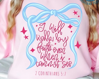 May include: A light pink sweatshirt with a light blue ribbon design and the text "I will walk by faith even when I cannot see" in red script. The text "2 Corinthians 5:7" is below the quote. Pink star accents surround the text.