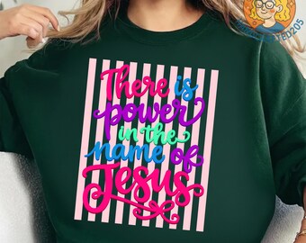 May include: Dark green sweatshirt with the text "There is power in the name of Jesus" in colorful script. The text is set against a background of pink and white vertical stripes. The sweatshirt has a crew neck and long sleeves.