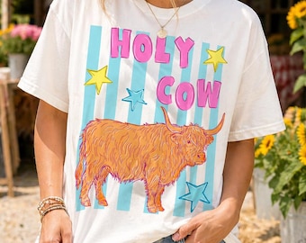 May include: Cream-colored t-shirt with the words "HOLY COW" in pink and blue, along with a cartoon illustration of a Highland cow. The design includes blue stripes, yellow stars, and blue stars.