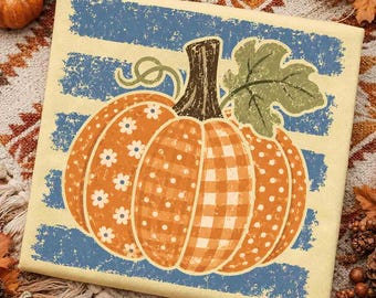 May include: A square canvas print featuring a central orange pumpkin with a dark brown stem and a green leaf. The pumpkin has sections with white floral, polka dot, and gingham patterns. The background consists of horizontal blue distressed stripes.