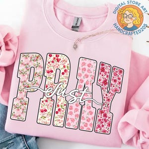 May include: A light pink sweatshirt with the word "PRAY" in large, floral-filled letters. The word "just" is written in a cursive font over the letters. The sweatshirt has a crew neck and long sleeves.