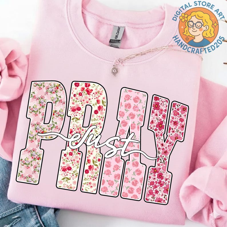 May include: A light pink sweatshirt with the word "PRAY" in large, floral-filled letters. The word "just" is written in a cursive font over the letters. The sweatshirt has a crew neck and long sleeves.