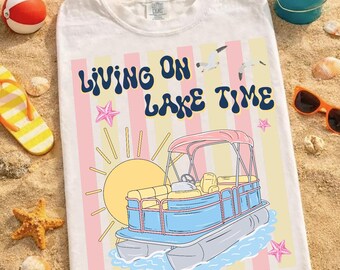 May include: White t-shirt with a colorful graphic design. The design features a pontoon boat, sun, seagulls, and the text "Living on Lake Time." The background has pink and yellow stripes, with starfish accents. Beach-themed apparel.