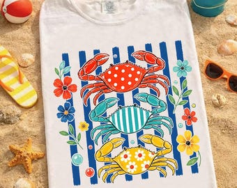 May include: White t-shirt featuring a colorful crab design. The design includes three crabs in red, teal, and yellow, each with a unique pattern, set against blue vertical stripes. Floral accents and a beach theme complete the design.