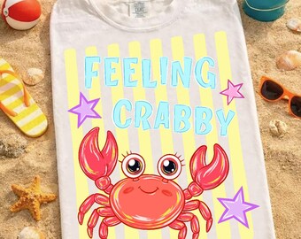 May include: White t-shirt with a cartoon crab design. The shirt features the text "FEELING CRABBY" in blue letters, with a crab illustration and star accents. The shirt is on a sandy surface with beach-themed accessories.