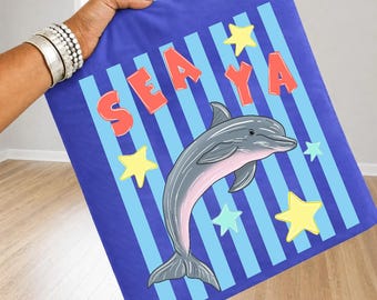 May include: A blue blanket with a dolphin illustration. The blanket features the word "SEA YA" in red letters, blue and light blue stripes, and yellow and light blue stars. The dolphin is gray with a pink belly.