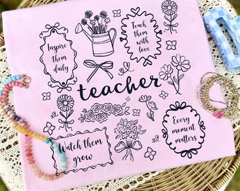 May include: A light pink t-shirt with the word "teacher" in a cursive font. The shirt features black line art of flowers, a watering can, and the phrases "Inspire them daily", "Teach them with love", "Watch them grow", and "Every moment matters".