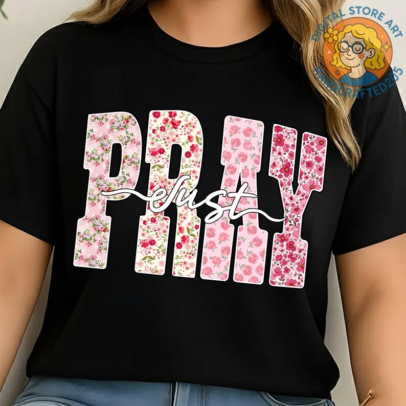 May include: Black t-shirt with the words "PRAY just" in a floral design. The letters are filled with pink and red flowers, and the word "just" is in a cursive font. The shirt has a round neckline and short sleeves.