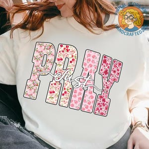 May include: Cream-colored sweatshirt featuring the word "PRAY" in large, block letters filled with pink floral patterns. The word "just" is written in a cursive font over the "PRAY" letters. A Digital Store Art and Handcrafted2023 logo is visible.