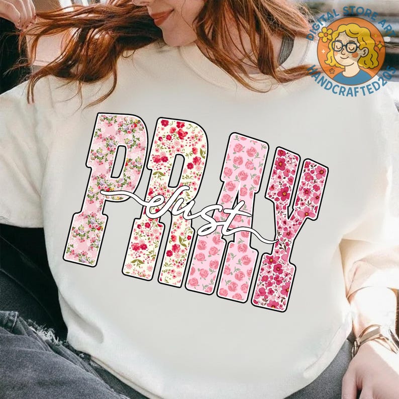 May include: Cream-colored sweatshirt featuring the word "PRAY" in large, block letters filled with pink floral patterns. The word "just" is written in a cursive font over the "PRAY" letters. A Digital Store Art and Handcrafted2023 logo is visible.