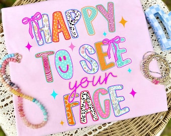 May include: A light pink t-shirt featuring the colorful text HAPPY TO SEE YOUR FACE in various patterns including leopard print, floral, and polka dots. The design includes pink bows, a smiley face, and small star icons printed on the fabric.