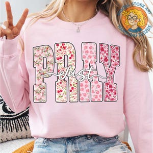 May include: A light pink sweatshirt with the words "PRAY just" in a floral design. The letters are filled with pink and red flowers. The word "just" is written in a cursive font over the letters.