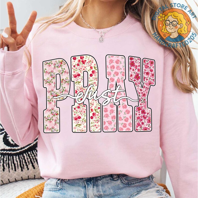 May include: A light pink sweatshirt with the words "PRAY just" in a floral design. The letters are filled with pink and red flowers. The word "just" is written in a cursive font over the letters.