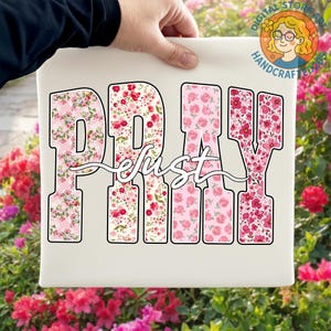 May include: A white canvas featuring the word "PRAY" in large, floral-patterned letters, with the word "just" in cursive script. The floral patterns include pink and red roses and other flowers. The canvas is held by a person.
