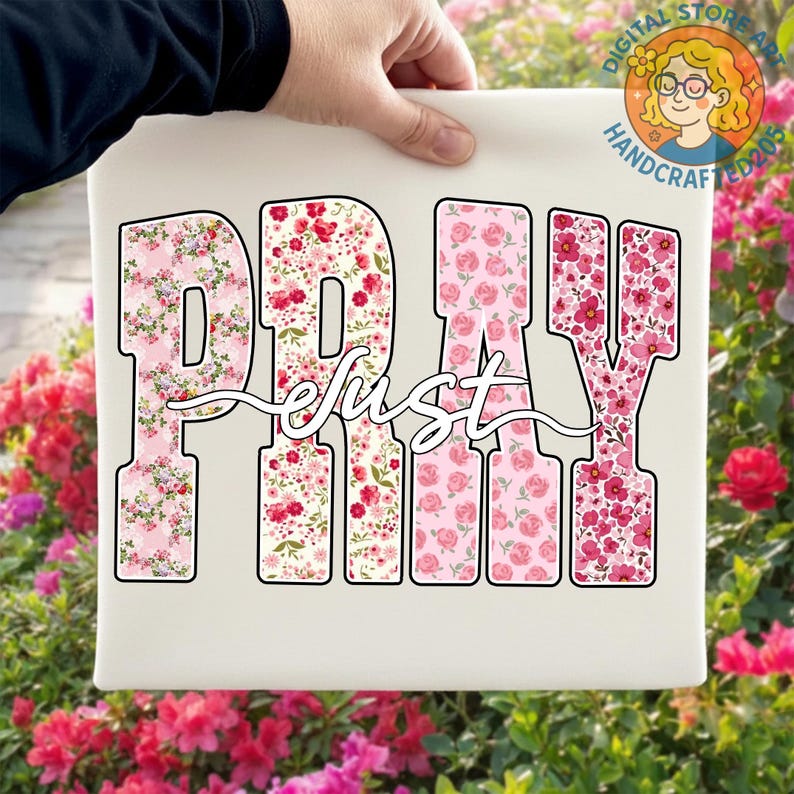 May include: A white canvas featuring the word "PRAY" in large, floral-patterned letters, with the word "just" in cursive script. The floral patterns include pink and red roses and other flowers. The canvas is held by a person.